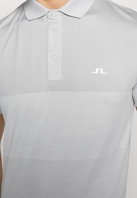 Ljust grå polo med tre knappar och en liten vit logotyp på vänster bröst, bärs av en person med armar delvis synliga.