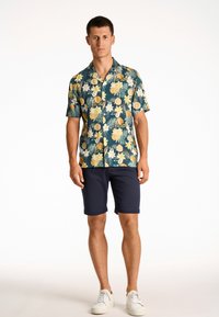 Camisa de flores de manga corta en azul marino con limones amarillos y flores blancas, combinada con pantalones cortos azul marino y zapatillas blancas. Tela suave, corte relajado.
