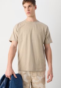 T-shirt en coton beige à manches courtes, affichant un design de logo subtil au centre. La personne porte une veste en denim dans une main.