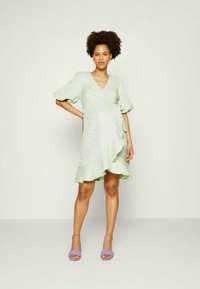 Vestido verde-claro com decote em V, bainha com babados e mangas curtas bufantes. Tecido texturizado e design assimétrico. Combinado com sandálias lavanda.