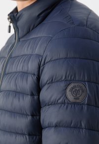 Marineblaue, isolierte Jacke mit gesteppter Optik, hohem Kragen, Reißverschluss und einem runden schwarzen Logo-Patch auf der linken Schulter.