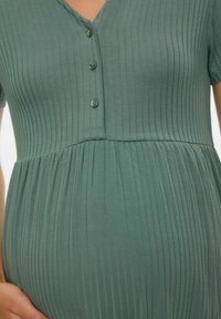 Robe de maternité verte à rayures avec des manches courtes, col en V et trois boutons décoratifs au niveau de la poitrine, fluide à partir d'un buste ajusté jusqu'à une jupe plissée.