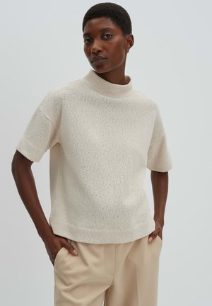 Vrouw met kort haar, gekleed in een crème kleurige gebreide coltrui met korte mouwen en beige broek, met handen in de zakken.