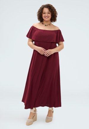 Smilende kvinde med krøllet hår iført en ærmeløs off-shoulder burgunder maxi-kjole og beige kilehæle med ankelsnørre.