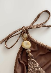 Brązowy top bikini z wzorem w węże. Posiada związany kokardę z teksturowanymi koralikami w odcieniach brązu i kremu w okolicy dekoltu.