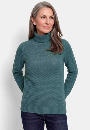 Vrouw met lang grijs haar, gekleed in een turquoise getextureerde coltrui en donkere spijkerbroek, staand met handen achter haar rug.