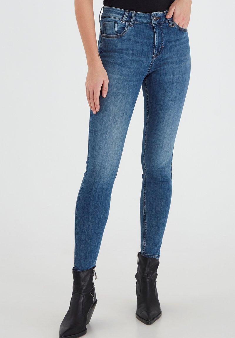 Tievās džinsēs vidēji zilos toņos, kas iekļauj klasisko piecu kabatu dizainu, jostas cilpiņas un rāvējslēdzēju, apvienotas ar melniem potīšu zābakiem.