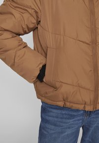 Brun puffjacka med dragkedja fram, med svarta ribbade ärmslut och sidofickor. Tyget är slätt och isolerat, vilket ger värme.