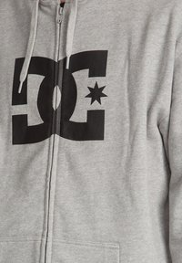 Haut à capuche zippé gris avec un grand logo DC noir et une étoile. Fabriqué en tissu doux avec une finition texturée et des cordons contrastés.