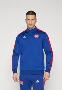 Blaue Adidas Trainingsjacke mit roten Streifen und Arsenal-Logo. Verfügt über einen hohen Kragen, einen durchgehenden Reißverschluss und gerippte Bündchen. Glattes Stoffgefühl.