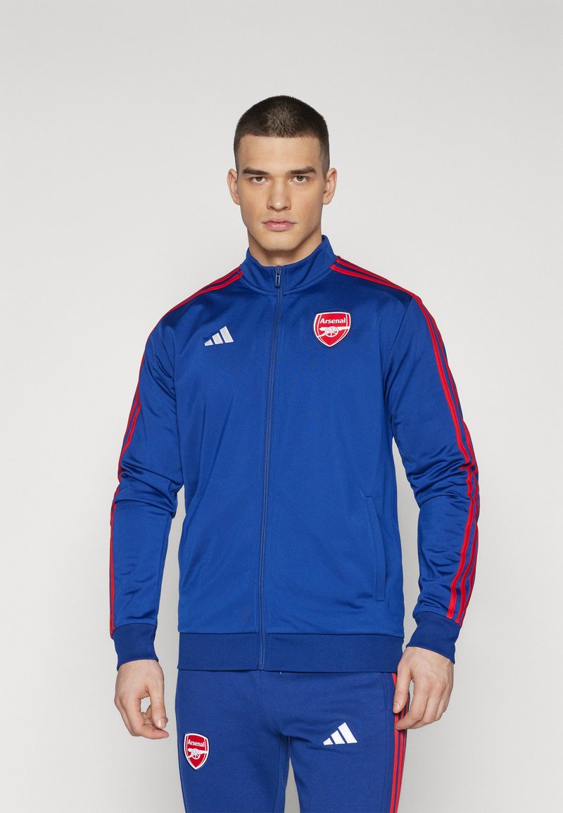 Blaue Adidas Trainingsjacke mit roten Streifen und Arsenal-Logo. Verfügt über einen hohen Kragen, einen durchgehenden Reißverschluss und gerippte Bündchen. Glattes Stoffgefühl.