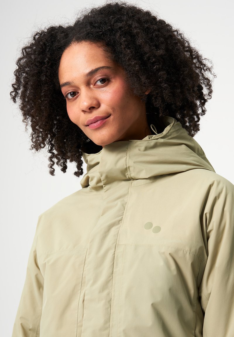 Helle beige wasserdichte Jacke mit hohem Kragen, strukturiertem Obermaterial, seitlichen Taschen und dezentem Branding. Mit Kapuze und Reißverschluss.