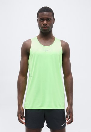 Atletyczny mężczyzna stoi, ubrany w jasnozielony bezrękawnik Nike i czarne krótkie spodenki Nike, zwrócony przodem na jednolitym jasnym tle.