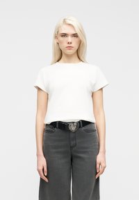 Jeune femme aux longs cheveux blonds portant un t-shirt blanc à manches courtes, un jean gris foncé et une ceinture noire avec une boucle en forme de cœur.