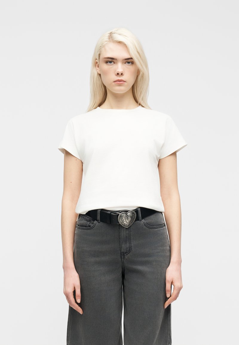 Jeune femme aux longs cheveux blonds portant un t-shirt blanc à manches courtes, un jean gris foncé et une ceinture noire avec une boucle en forme de cœur.