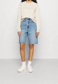 Sweatshirt beige avec texte rose, shorts en denim bleu clair et baskets blanches avec accents jaunes. Fond clair, position debout.