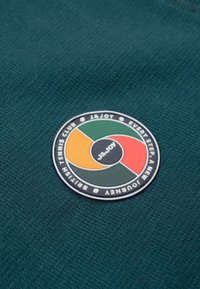 Groene textuurstof met een cirkelvormig, veelkleurig logo waarop "J&JOY" en "BRITISH TENNIS CLUB" rond de rand is staat.