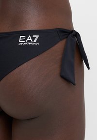 Svart bikinitruse med knyting på den ene siden, med en hvit EA7 Emporio Armani-logo på midjebåndet. Glatt tekstur, minimalistisk design.