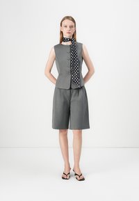 Gilet grigio senza maniche con bottoni, abbinato a pantaloni corti grigi. Accentuato con un foulard a pois bianco e nero. Indossato con infradito nere.