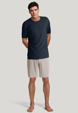 Marineblaue Kurzarmbluse mit glatter Textur, kombiniert mit hellen, gemusterten Shorts mit einem Diamantdesign, präsentiert an einem stehenden Modell.
