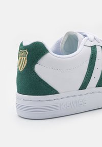 Bílé tenisky s zelenými semišovými akcenty, kulatou špičkou, plochou podrážkou a vyšitým logem na patě; obsahují kontrastní textury a švy.