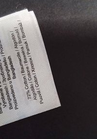 Etiqueta de tela blanca, con texto impreso que indica 72% algodón en varios idiomas, con un borde ligeramente deshilachado sobre un fondo negro.