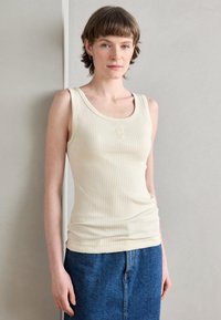 Top in crema a costine con spalline larghe e scollatura a barca; presenta un logo goffrato discreto sul petto. Abbinato a una gonna di denim.