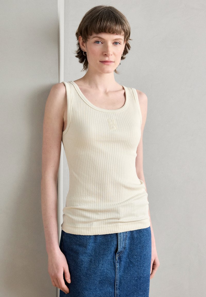 Top in crema a costine con spalline larghe e scollatura a barca; presenta un logo goffrato discreto sul petto. Abbinato a una gonna di denim.