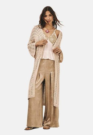 Kimono de encaje beige sobre una blusa blanca ajustada con volantes. Pantalones anchos beige con brillo, complementados por un collar llamativo.