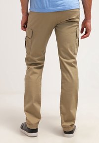 Pantalones cargo beige con bolsillos laterales, piernas rectas y una textura suave. Llevados con zapatillas negras sobre un fondo neutro.