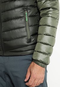 Puhvi takki, millel on tume roheline keha ja heledamat roheline varrukad, millega on horisontaalsed tikitud sektsioonid ja lukuga tasku roheline tõmblukk.