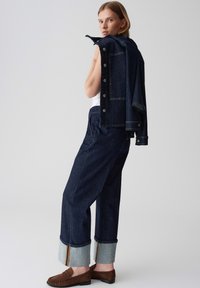 Veste en denim foncé avec des boutons aux épaules, associée à un jean foncé taille haute avec des poignets en denim clair et des mocassins marron.