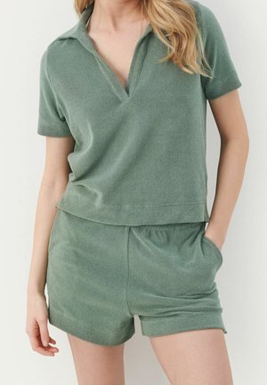 Kobieta ubrana w miękki zielony top z krótkim rękawem i kołnierzykiem oraz dopasowane szorty, trzymająca lewą rękę w kieszeni na jednolitym tle.