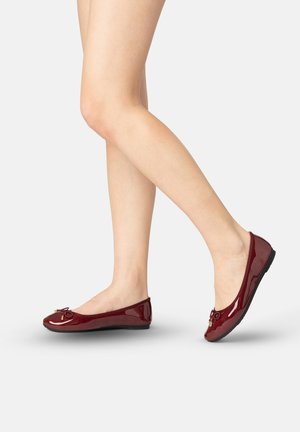 Jambes portant des ballerines brillantes bordeaux avec de petits nœuds, un pied à plat et l'autre légèrement levé sur un fond clair.