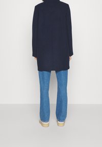 Mörkblå ullkappa med högt krage, rak passform och långa ärmar; kombinerad med ljusblå utsvängda jeans och tan plattformsskor.