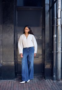 Hvid tekstureret jakke med lynlås og oversize ærmer, kombineret med blå vidde ben cowboybukser og hvide spidse hæle.