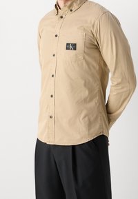 Světle hnědá košile na knoflíky s náprsní kapsou, s černým logem a tmavými knoflíky. Hladký materiál, klasický límec a dlouhé rukávy.