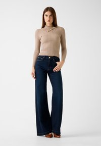 Haut en maille beige côtelée avec des boutons aux épaules, associé à un jean bleu foncé à jambes larges. Chaussures plates marron complètent la tenue.