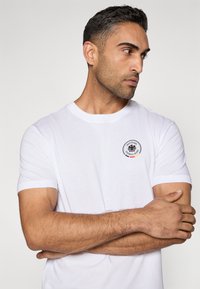 Balta kokvilnas T-krekls ar apaļu kaklu, īsām piedurknēm un aplī izcelta melna ērglis ar vācu tekstu.