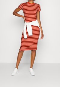 Červené a bílé pruhované šaty s přiléhavým siluetou a krátkými rukávy, spárované s uvázanou bílou mikinou a bílými teniskami.