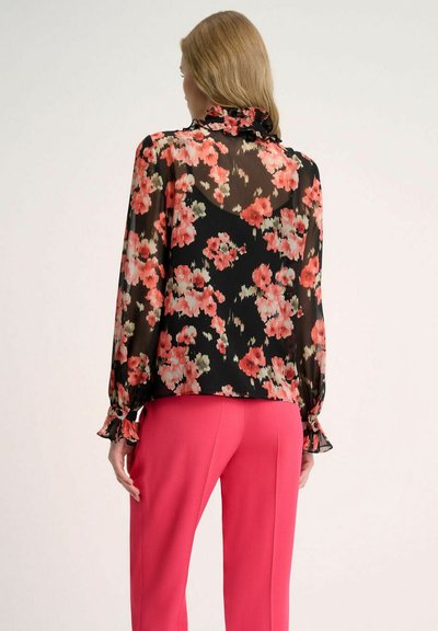 Blusa negra con estampado floral en tonos rosas y verdes, con escote volado, mangas largas y puños fruncidos, combinada con pantalones rosas.