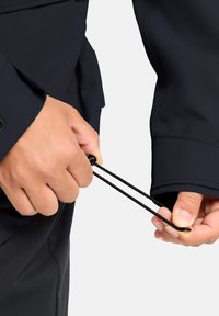 Giacca blu navy con regolazione a cordoncino elastico ai polsini, caratterizzata da una texture liscia e un design aderente. Mani che stringono il cordoncino per stringere.