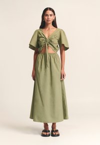 Dvo-delna obleka olivno zelene barve z izrezanim zgornjim delom, ki ima sprednjo zavezavo in rahlo razširjene rokave, v kombinaciji z usklajeno dolgo krilo.