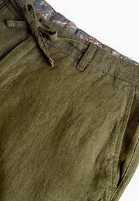 Pantalons olive avec passants de ceinture, une ceinture en tissu nouée, un revêtement de taille à motifs floraux et des poches latérales sur un fond blanc.