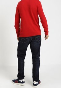 Roter Strickpullover mit strukturiertem Muster, kombiniert mit dunkelblauen Jeans, die hinten Taschen mit einem markanten Emblem haben. Sneaker sind sichtbar.