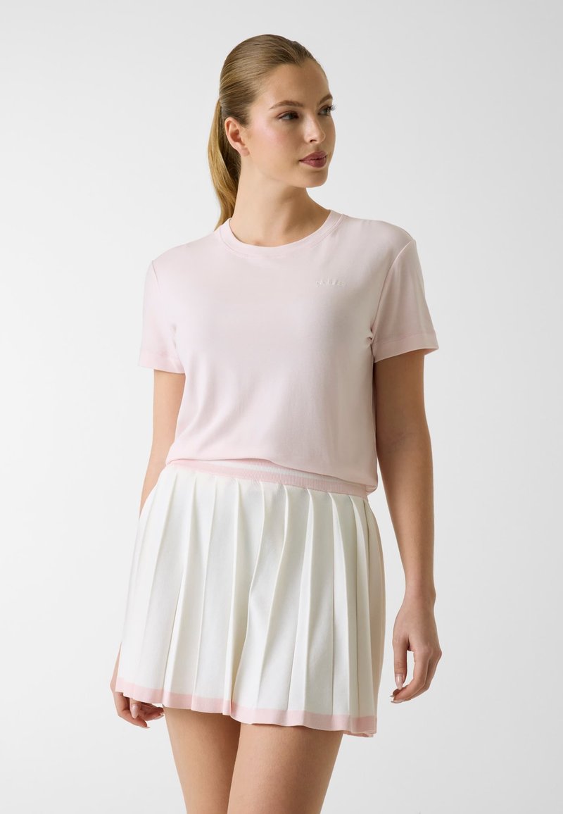 Jeune femme avec une queue de cheval portant un t-shirt rose clair à manches courtes et une jupe plissée blanche avec un ourlet rose, debout devant un fond uni.