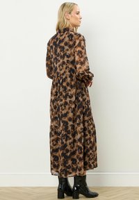 Kobieta z długimi blond włosami, ubrana w midi sukienkę z długim rękawem w brązowo-czarny wzór oraz czarne lakierowane botki do kostek, stojąca bokiem.