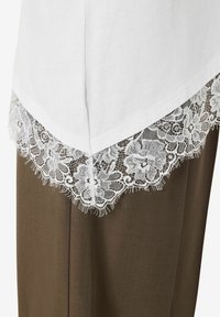 Hvit t-skjorte med V-formet kant, med en delikat hvit blonderkant nederst. Blonderne har blomsteraktige mønstre og frynse detaljer.