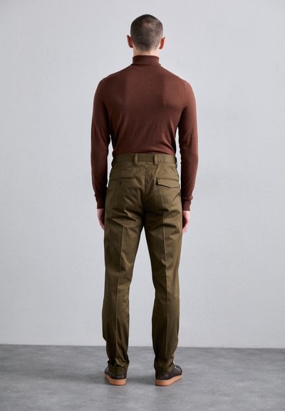 Brūns pieguļošs turtlenecks krekliņš saskaņots ar olīvzaļiem biksēm. Biksēm ir jostas cilpas, divas aizmugurējās kabatas un sašaurinātas kājas. Valkā brūnas kurpes.