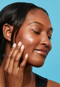 Hydratisierendes Hautpflegeprodukt, das auf strahlend brauner Haut aufgetragen wird. Die Person hat lange schwarze Haare mit glatter Textur und zarten rosa Nägeln.